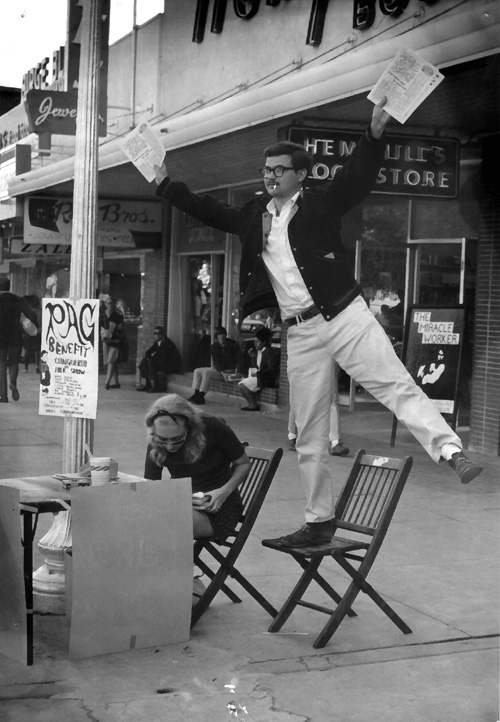 George Vizard balances on one foot to hawk the second issue of The Rag on the Drag. His companion, Mariann Vizard, sits nearby.